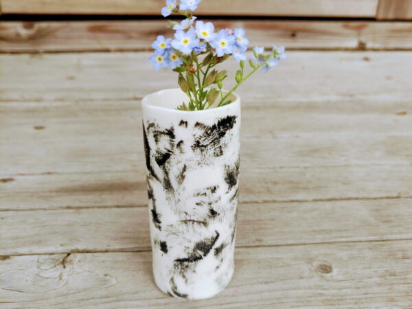 The Rustic Elegance Porcelain Mini Vase, designed in white with black patterns, holds a bunch of tiny blue flowers on a wooden surface. The Rustic Elegance Porcelain Mini Vase, designed in white with black patterns, holds a bunch of tiny blue flowers on a wooden surface.