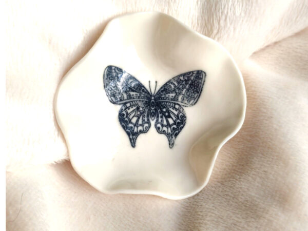 A white ceramic Butterfly Ring Holder (7cm x 7cm) with irregular edges and a black butterfly illustration in the center is placed on a soft white fabric.