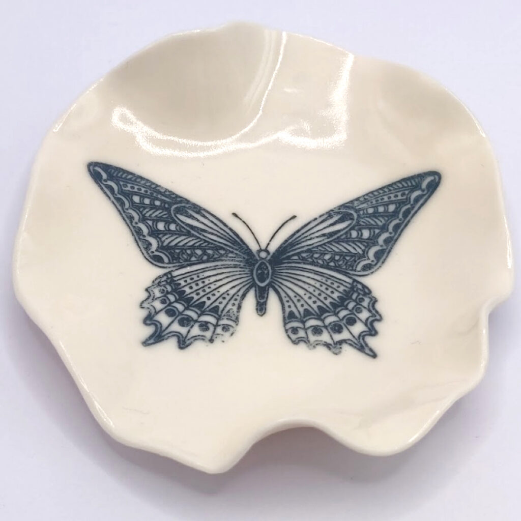A small, cream-colored ceramic dish with a detailed black butterfly illustration at the center.
