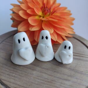 The Love Ghost Trio, featuring three small white clay ghost figures with black eyes and heart details, is showcased in front of an orange flower on a wooden surface. The Love Ghost Trio, featuring three small white clay ghost figures with black eyes and heart details, is showcased in front of an orange flower on a wooden surface.