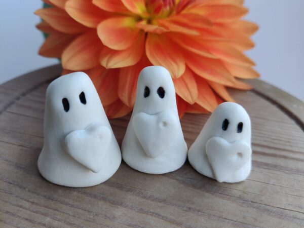 The Love Ghost Trio, featuring three small white clay ghost figures with black eyes and heart details, is showcased in front of an orange flower on a wooden surface. The Love Ghost Trio, featuring three small white clay ghost figures with black eyes and heart details, is showcased in front of an orange flower on a wooden surface.