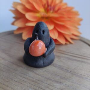 The Midnight Mini Ghost Figurine 🎃, a small black clay figure holding an orange pumpkin, is placed on a wooden surface with a large orange flower in the background. The Midnight Mini Ghost Figurine 🎃, a small black clay figure holding an orange pumpkin, is placed on a wooden surface with a large orange flower in the background.