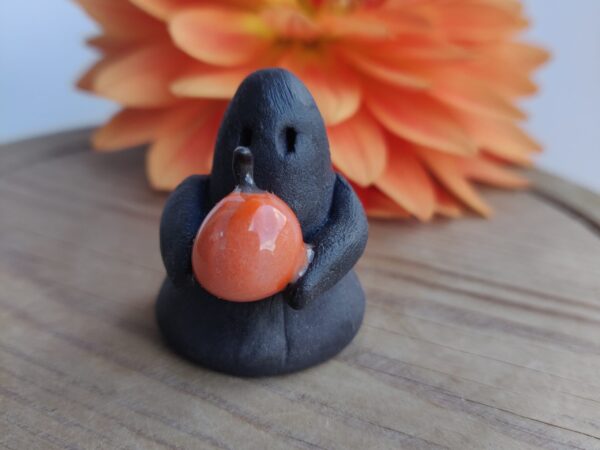 The Midnight Mini Ghost Figurine 🎃, a small black clay figure holding an orange pumpkin, is placed on a wooden surface with a large orange flower in the background. The Midnight Mini Ghost Figurine 🎃, a small black clay figure holding an orange pumpkin, is placed on a wooden surface with a large orange flower in the background.