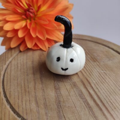 A Cheeky Porcelain Pumpkin, featuring a cute face and a small white exterior, rests on a wooden surface with an orange flower visible in the background. A Cheeky Porcelain Pumpkin, featuring a cute face and a small white exterior, rests on a wooden surface with an orange flower visible in the background.