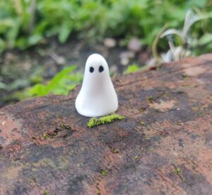 A piece from the Mini Ghostly Gathering Porcelain Collection, featuring a small, smooth white figurine with two black dots for eyes, is displayed on a moss-covered brick outdoors.