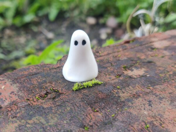A piece from the Mini Ghostly Gathering Porcelain Collection, featuring a small, smooth white figurine with two black dots for eyes, is displayed on a moss-covered brick outdoors. A piece from the Mini Ghostly Gathering Porcelain Collection, featuring a small, smooth white figurine with two black dots for eyes, is displayed on a moss-covered brick outdoors.