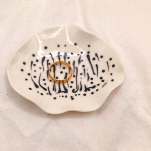 A gold ring rests on the Squiggles and Dots porcelain ring holder, featuring black dotted and striped patterns, placed on a white fabric background. A gold ring rests on the Squiggles and Dots porcelain ring holder, featuring black dotted and striped patterns, placed on a white fabric background.
