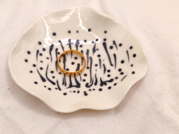A gold ring rests on the Squiggles and Dots porcelain ring holder, featuring black dotted and striped patterns, placed on a white fabric background.