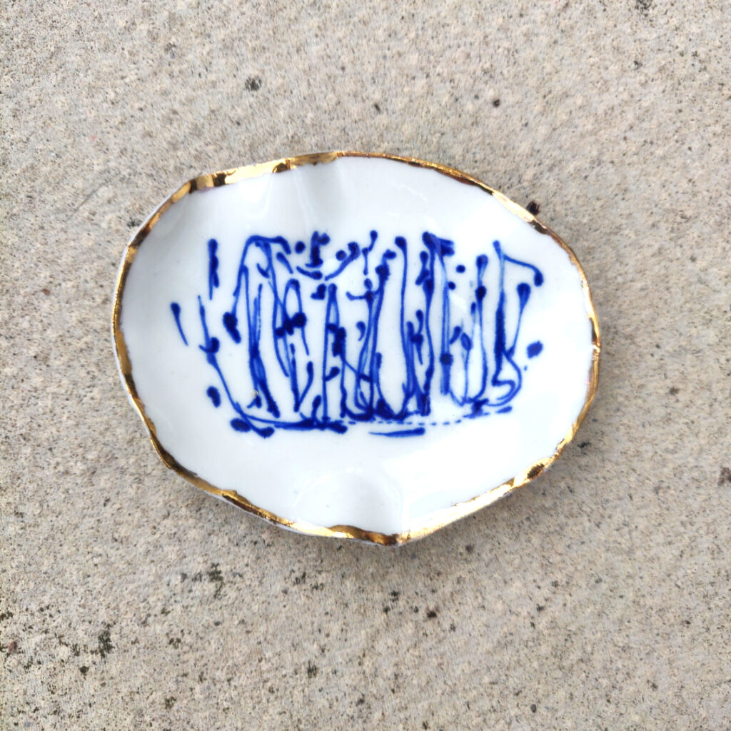 The Blue and White Porcelain Ring Holder with gold lustre features a white base adorned with abstract blue patterns and is elegantly finished with gold-trimmed edges, all set against a textured gray surface.
