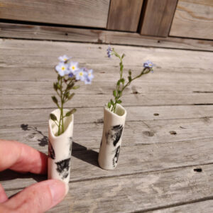 Two Ink-Stroke Miniature Porcelain Vases, featuring black markings on a white cylindrical surface and each containing delicate purple flowers, rest on a wooden surface as a hand carefully adjusts one of them. Two Ink-Stroke Miniature Porcelain Vases, featuring black markings on a white cylindrical surface and each containing delicate purple flowers, rest on a wooden surface as a hand carefully adjusts one of them.