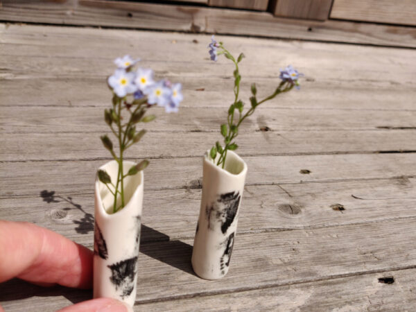 Two Ink-Stroke Miniature Porcelain Vases, featuring black markings on a white cylindrical surface and each containing delicate purple flowers, rest on a wooden surface as a hand carefully adjusts one of them. Two Ink-Stroke Miniature Porcelain Vases, featuring black markings on a white cylindrical surface and each containing delicate purple flowers, rest on a wooden surface as a hand carefully adjusts one of them.