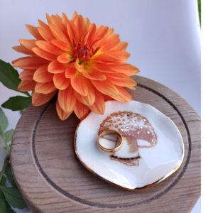 An orange flower is positioned beside the Enchanted Forest Porcelain Mushroom Ring Holder/Trinket with Gold Lustre, which holds two gold rings and rests on a round wooden surface. An orange flower is positioned beside the Enchanted Forest Porcelain Mushroom Ring Holder/Trinket with Gold Lustre, which holds two gold rings and rests on a round wooden surface.