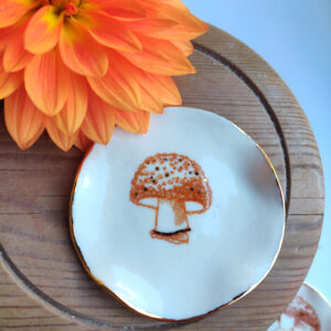 An orange flower is set beside the Enchanted Forest Porcelain Mushroom Ring Holder/Trinket with Gold Lustre, elegantly placed on a wooden surface. An orange flower is set beside the Enchanted Forest Porcelain Mushroom Ring Holder/Trinket with Gold Lustre, elegantly placed on a wooden surface.