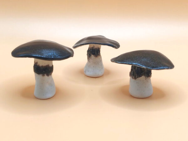 Three ceramic mushrooms with dark caps and light stems are arranged on a beige surface. Three ceramic mushrooms with dark caps and light stems are arranged on a beige surface.