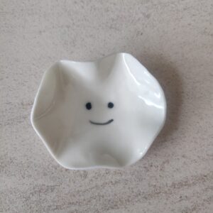 A compact, white ceramic dish featuring a wavy edge and adorned with a minimalist smiley face design at its base, known as the Mini Smiley Face. A compact, white ceramic dish featuring a wavy edge and adorned with a minimalist smiley face design at its base, known as the Mini Smiley Face.