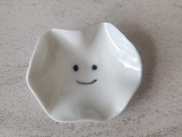 A compact, white ceramic dish featuring a wavy edge and adorned with a minimalist smiley face design at its base, known as the Mini Smiley Face. A compact, white ceramic dish featuring a wavy edge and adorned with a minimalist smiley face design at its base, known as the Mini Smiley Face.