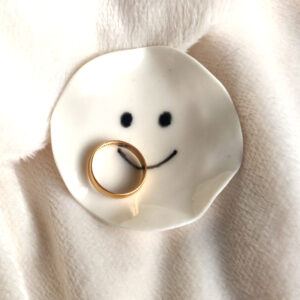 The Smiling Face Ring Holder, a small ceramic dish with a drawn smiley face, holds a gold ring and is placed on a soft, light-colored fabric. The Smiling Face Ring Holder, a small ceramic dish with a drawn smiley face, holds a gold ring and is placed on a soft, light-colored fabric.