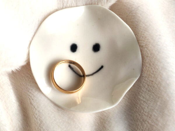 The Smiling Face Ring Holder, a small ceramic dish with a drawn smiley face, holds a gold ring and is placed on a soft, light-colored fabric.